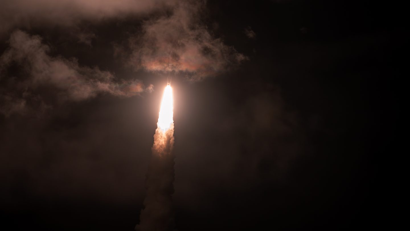 An Atlas V rocket's bright exhaust flame climbing through orange-lit clouds in the night sky after launch