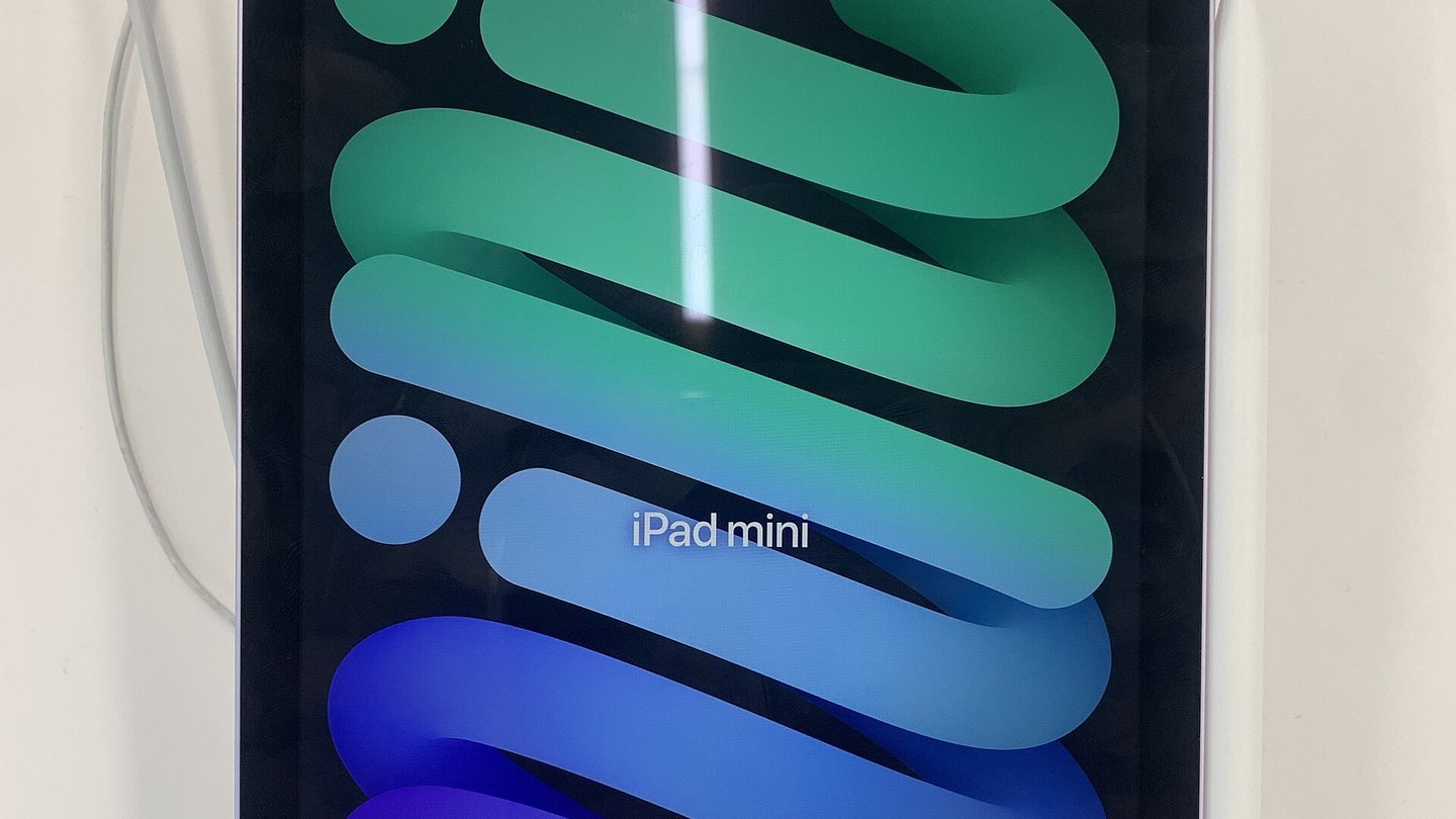 The screen of an iPad mini showing the words 'iPad mini' over green, blue and purple curved stripes, standing on a white surface