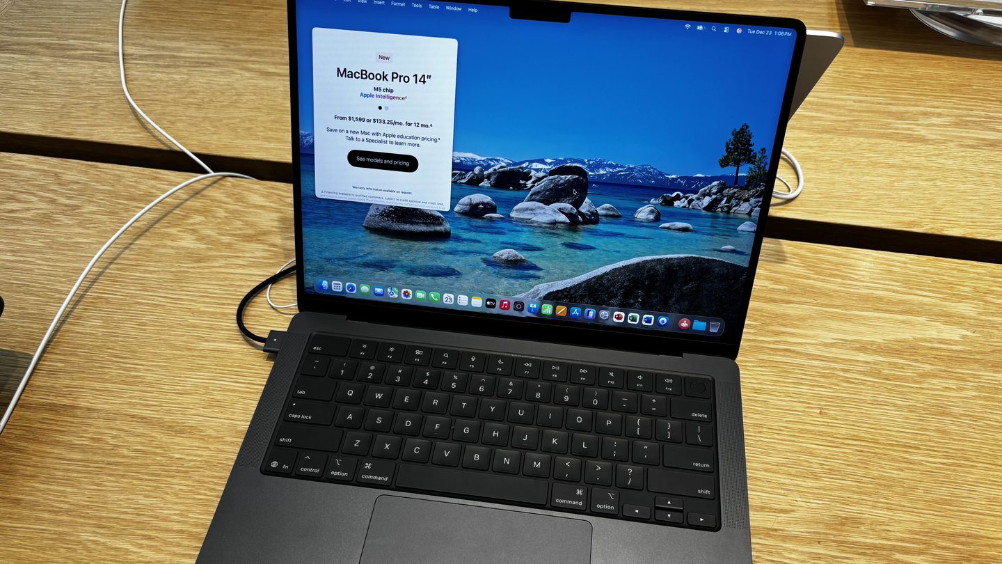 A space black 14-inch MacBook Pro laptop open on a wooden store table, its screen showing a lake landscape and a 'MacBook Pro 14-inch' information panel