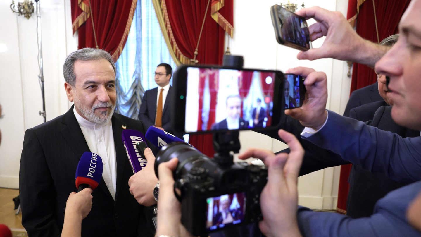 Iranian Foreign Minister Abbas Araghchi, in a dark suit, speaks to reporters holding microphones and phones in a room with red curtains