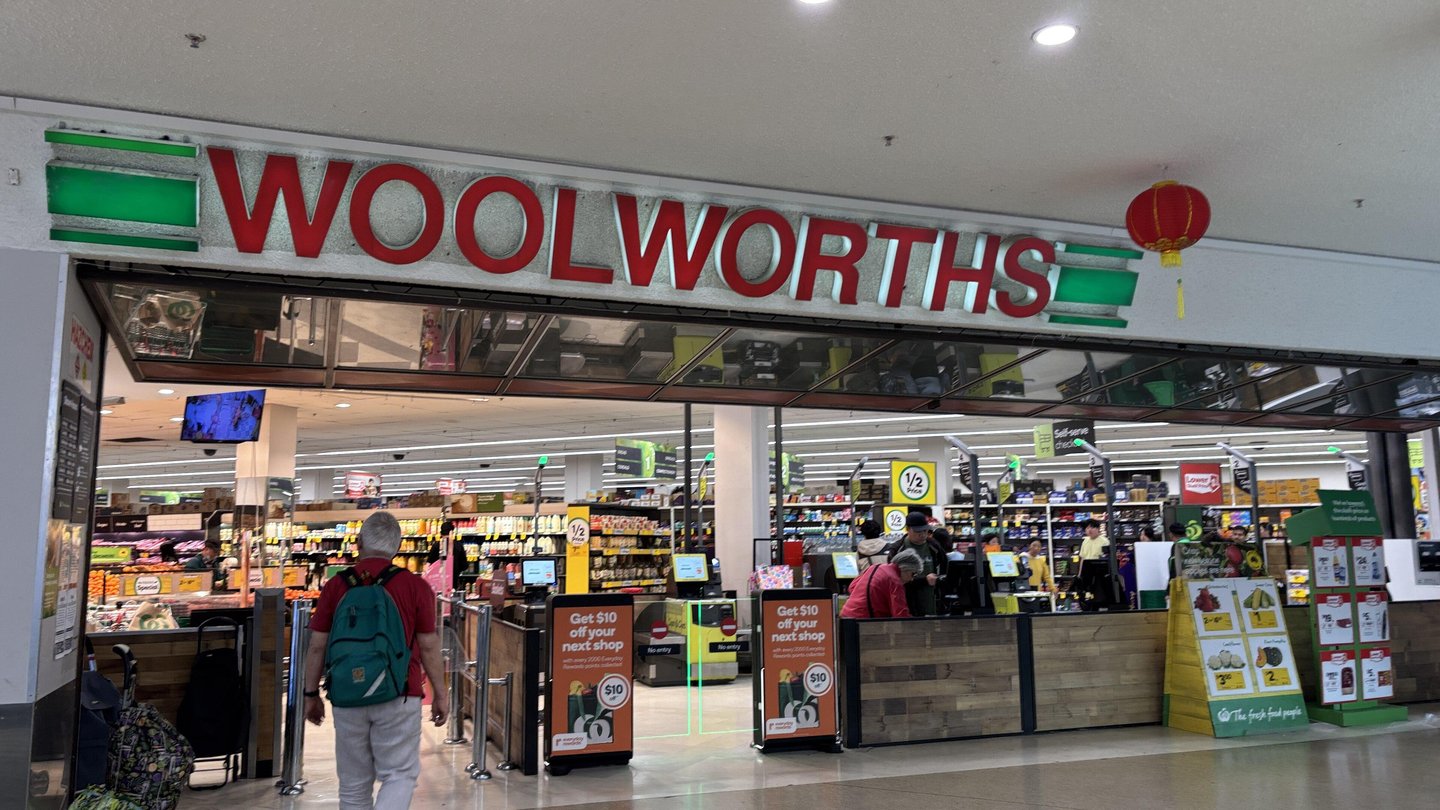 The entrance and checkouts of a Woolworths supermarket inside a shopping centre, with a shopper walking in