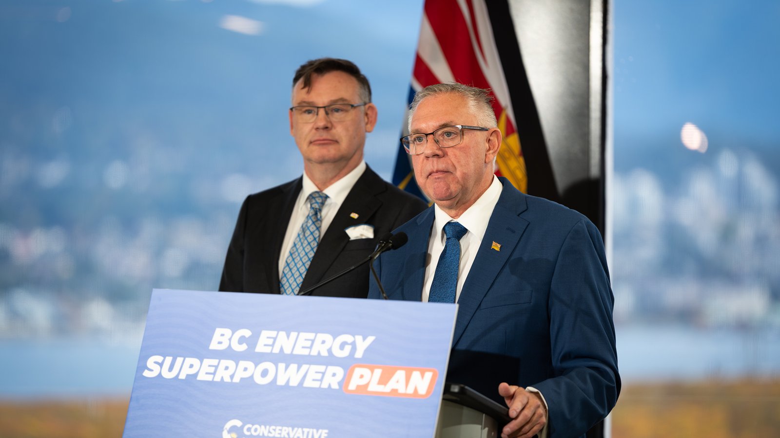 BC Conservative leader Lorne Doerkson, in glasses and a blue suit, speaks at a lectern reading 'BC Energy Superpower Plan', with another man and the British Columbia flag behind him