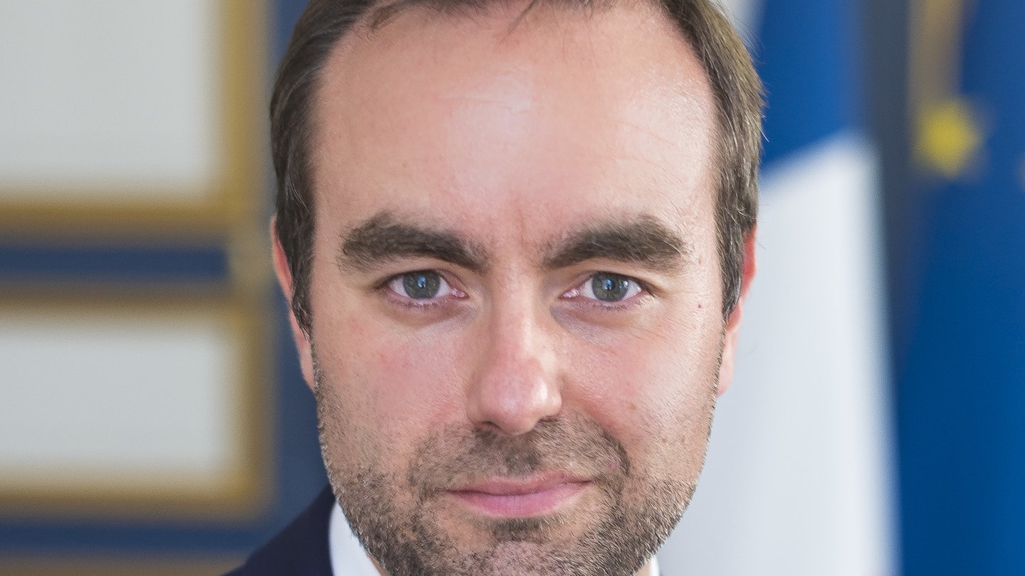 Portrait of Sébastien Lecornu in a dark suit and blue tie in front of French and European flags
