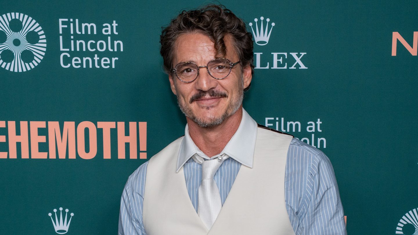 Pedro Pascal in glasses, a cream waistcoat and a pale striped shirt smiling in front of a green Behemoth! backdrop with Film at Lincoln Center logos