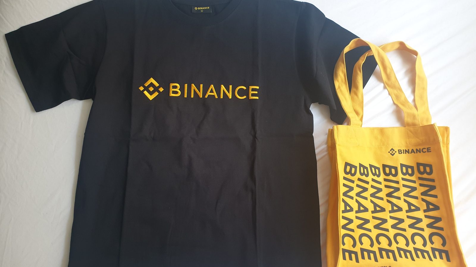 A black T-shirt with the yellow Binance logo next to a yellow Binance tote bag