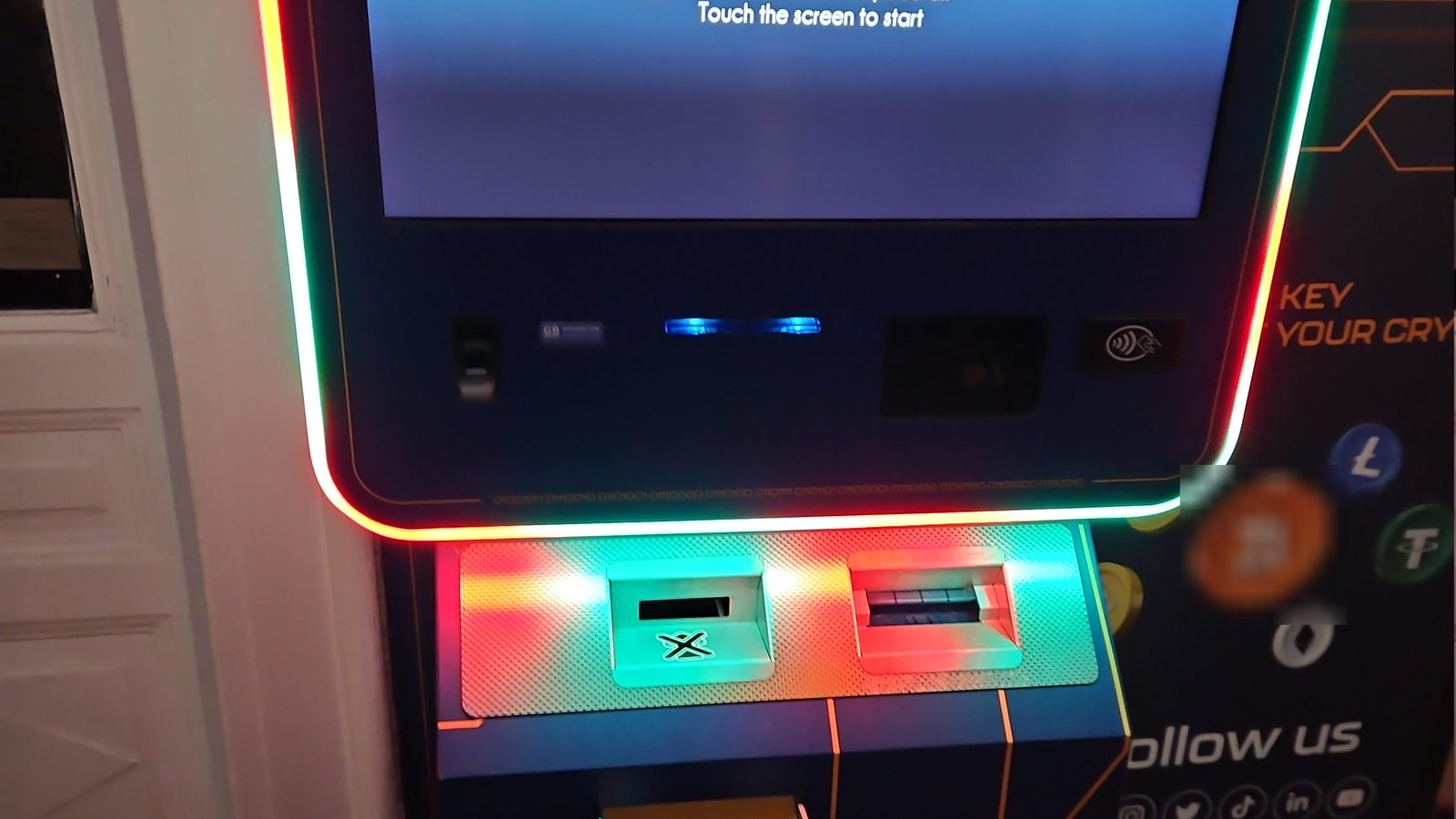 A cryptocurrency ATM with coloured lights and crypto logos at a railway station
