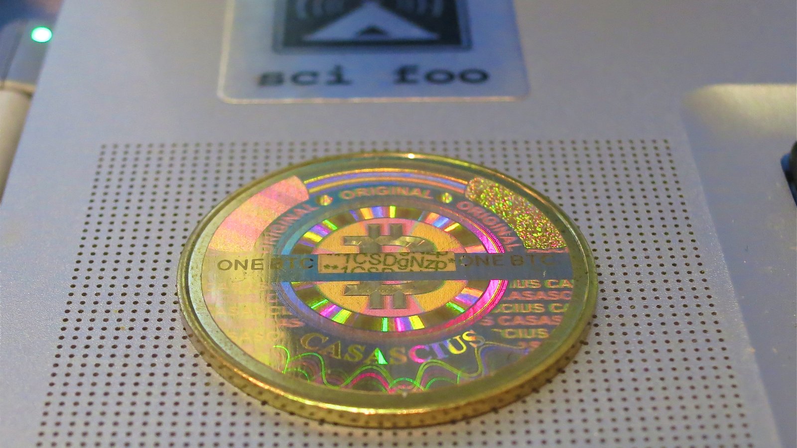 A gold-coloured physical bitcoin coin with a rainbow hologram sticker lying on a perforated white surface