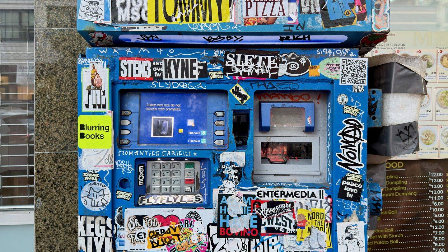 A public bitcoin ATM covered in stickers and graffiti tags, with its screen offering a bitcoin option