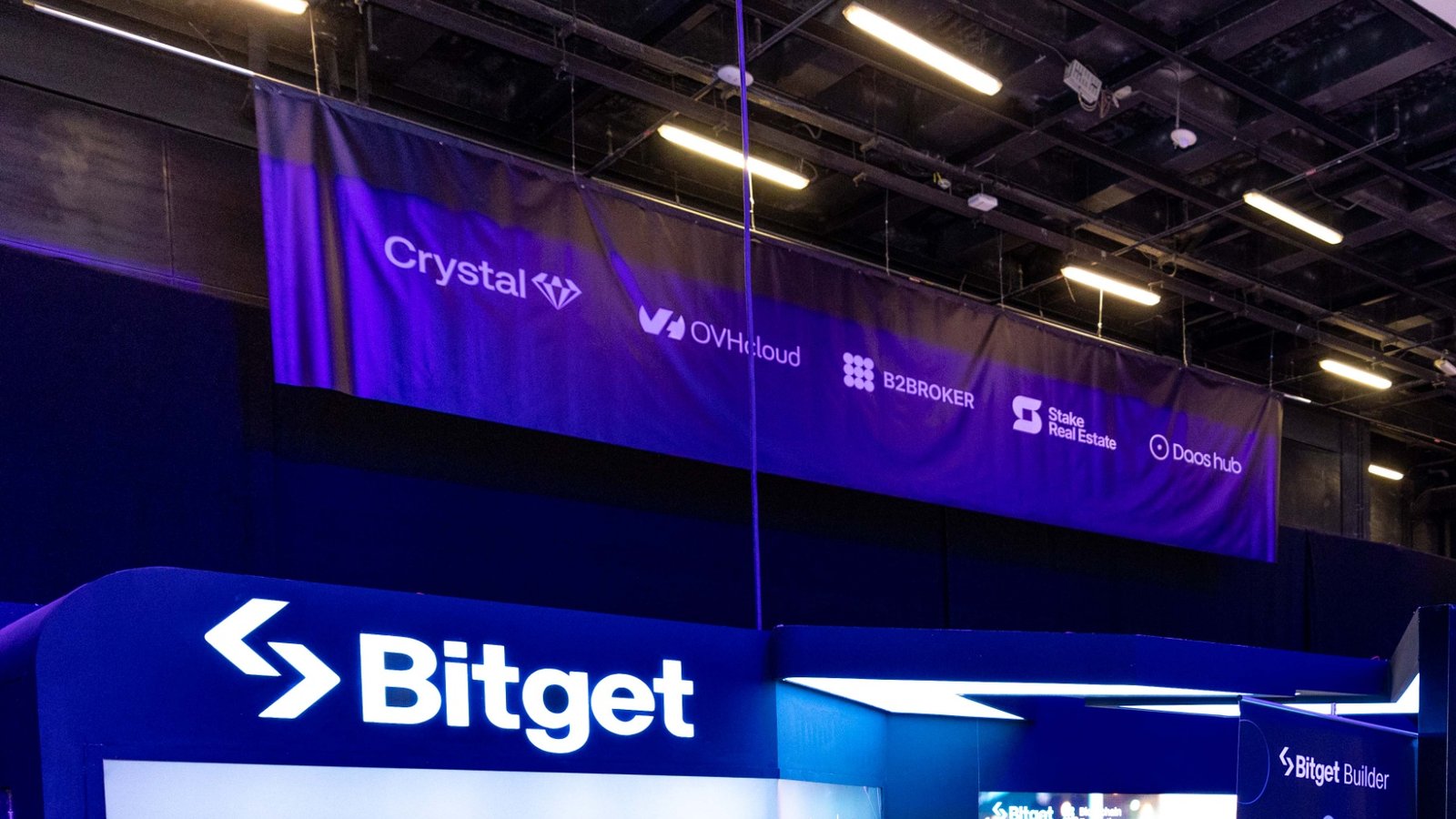 Illuminated Bitget logo on an exhibition stand beneath a sponsor banner in a conference hall