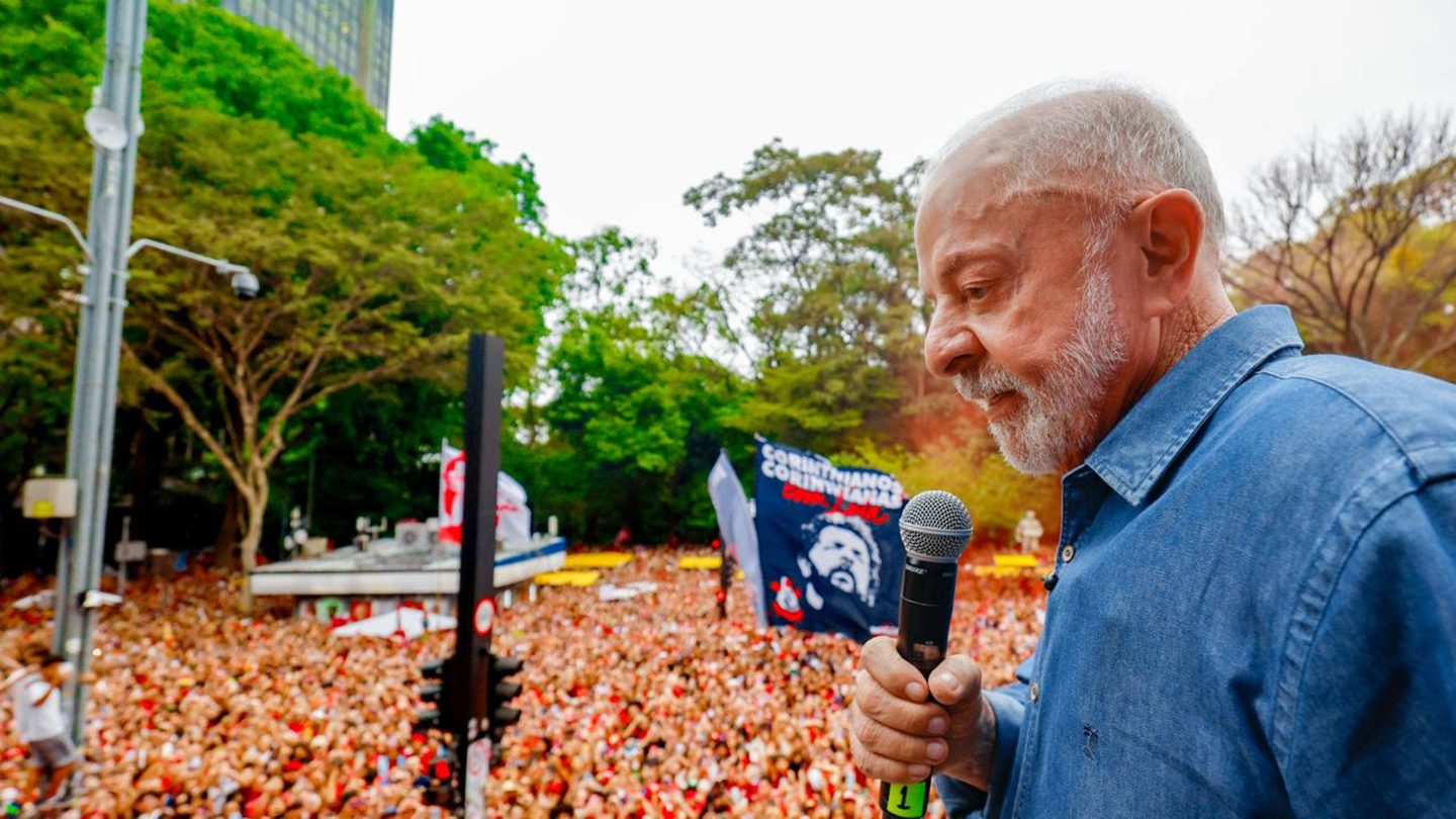 Luiz Inácio Lula da Silva, in a blue denim shirt, speaks into a microphone above a large crowd on a tree-lined avenue, with a banner bearing his image in the background