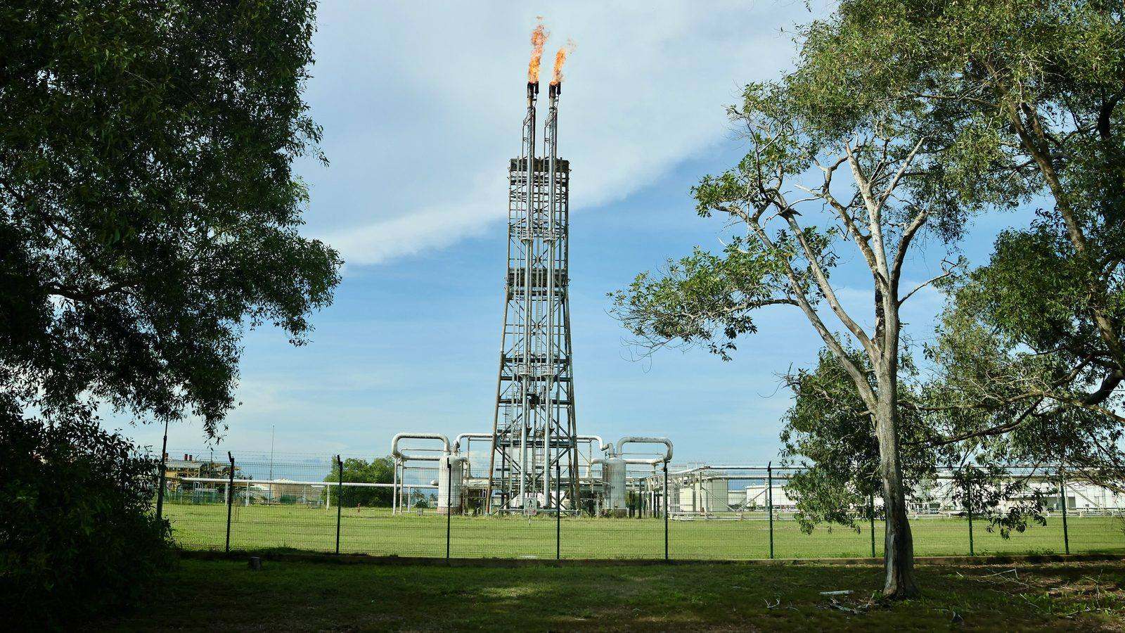 A tall steel flare stack burning two small flames at an oil refinery, behind a fence and lawn
