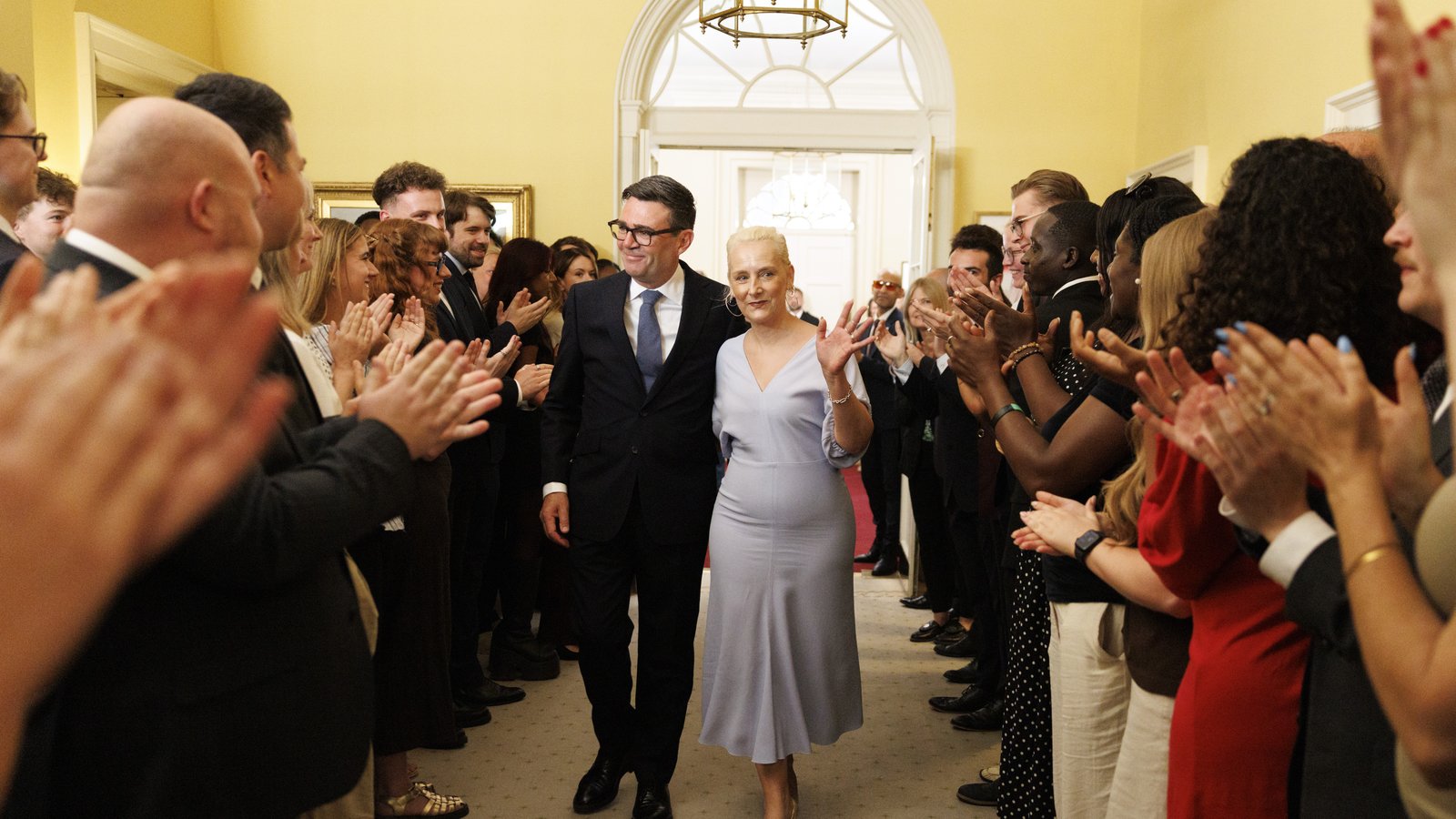 Andy Burnham, in a dark suit and glasses, walks with his wife through a corridor of applauding staff inside 10 Downing Street
