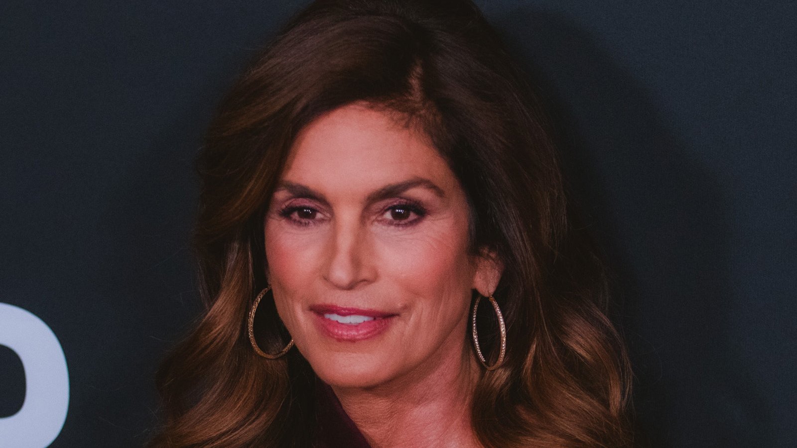 Cindy Crawford, with long brown hair and hoop earrings, smiles in front of a dark background