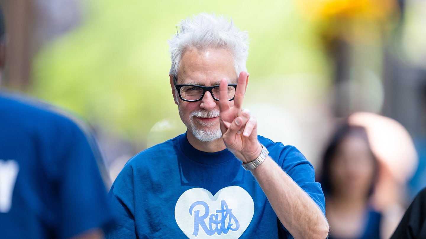 Film-maker James Gunn, with white hair, glasses and a blue T-shirt, points a finger upward on a city street