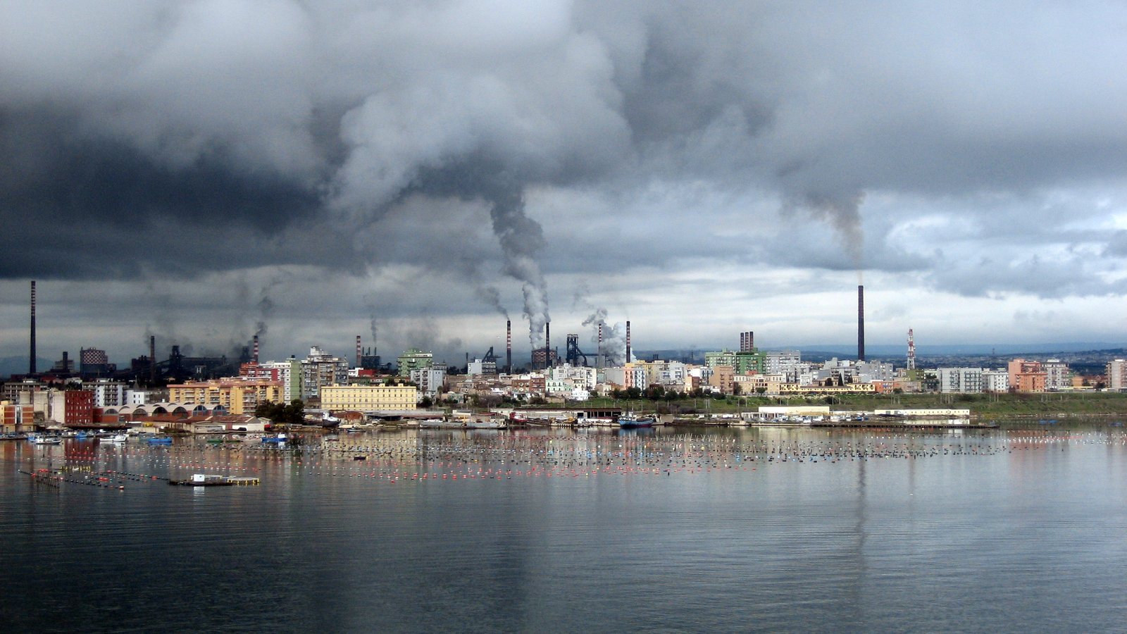 The ILVA steelworks in Taranto seen across open ground, with chimneys and industrial buildings