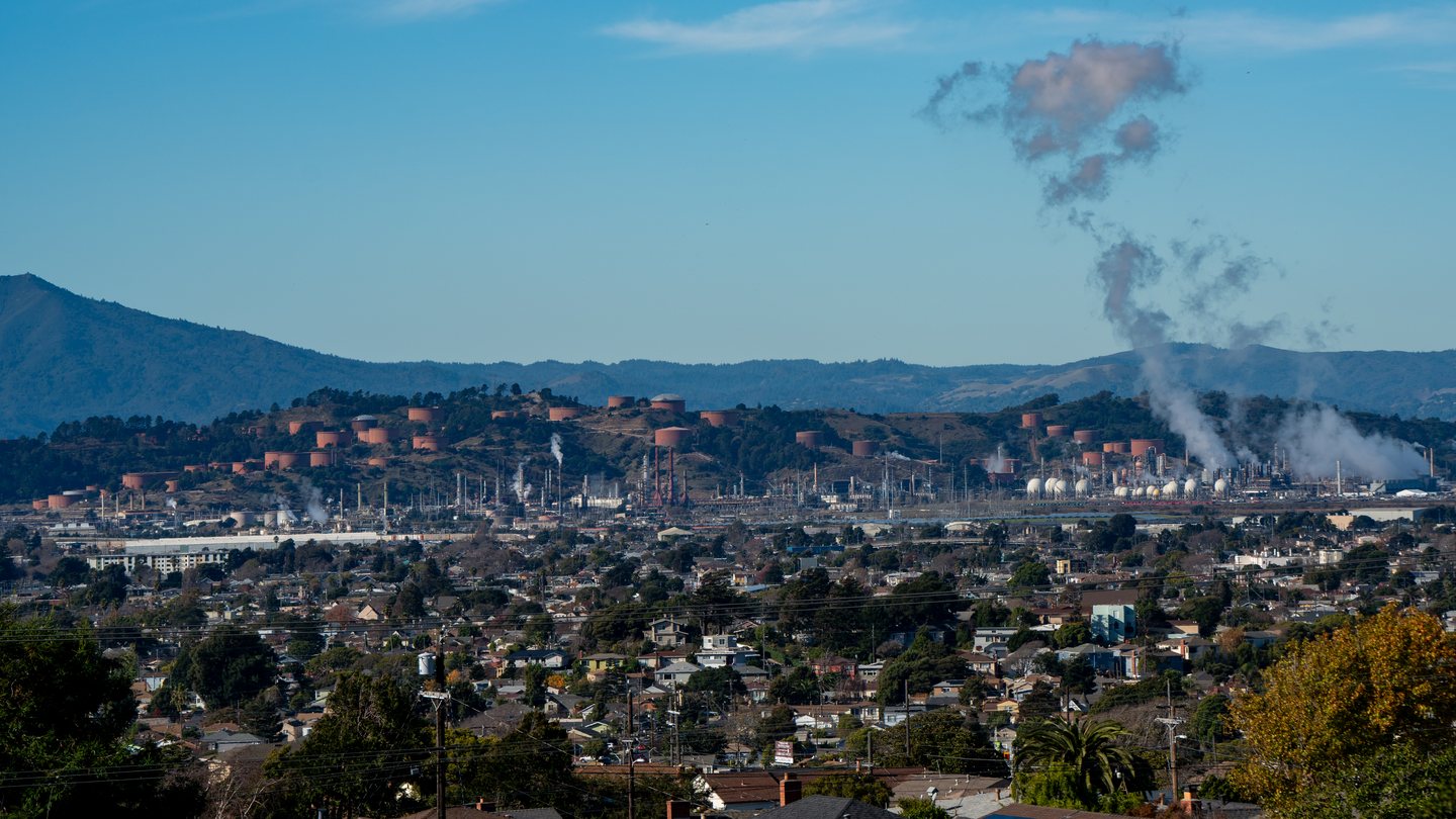 Chevron's Richmond oil refinery in California, with steam rising from its stacks above rows of houses and hills behind
