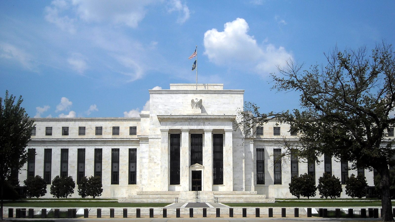 The white marble Marriner S. Eccles Federal Reserve Board Building in Washington under a blue sky, with trees in front