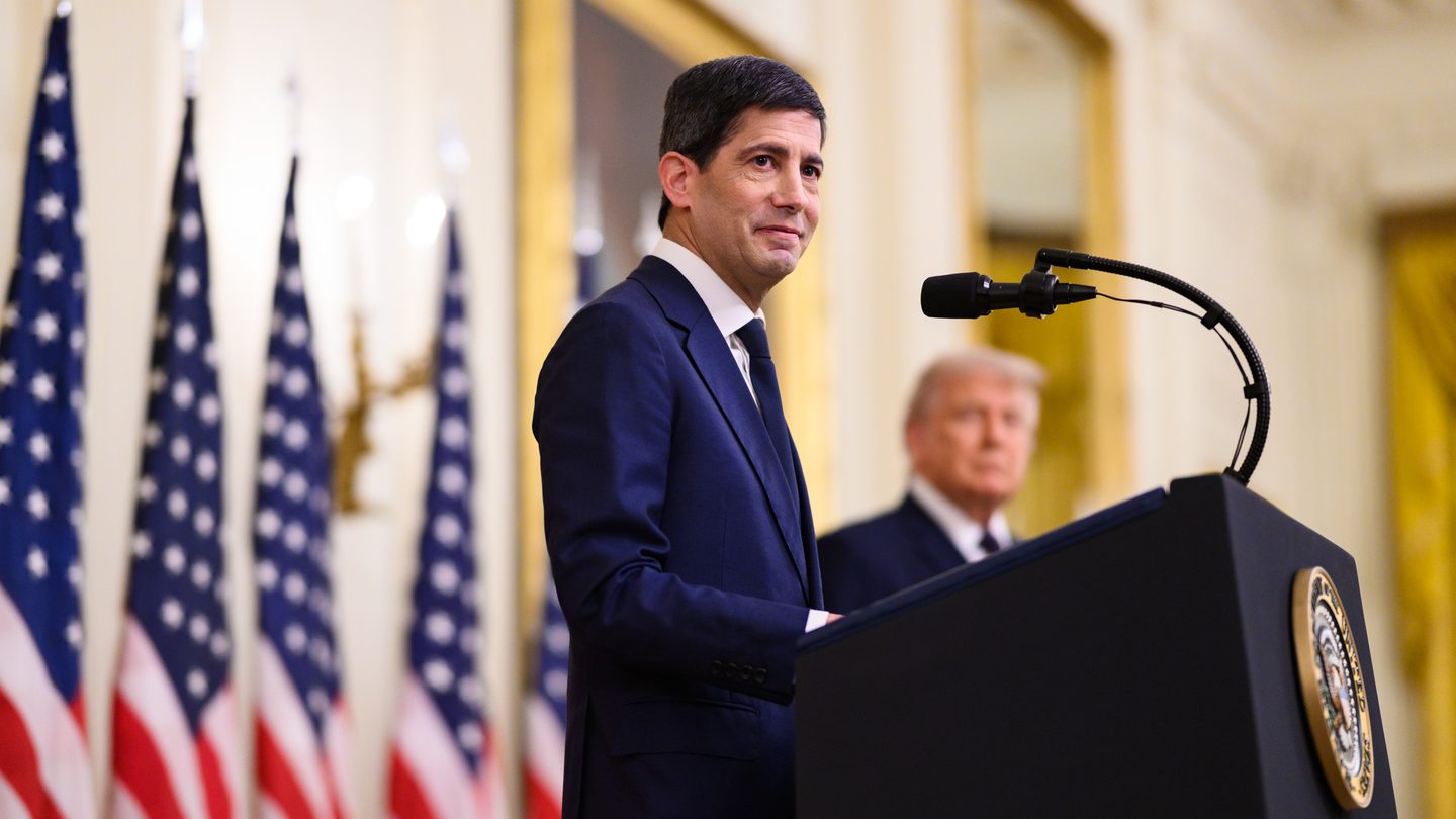 Federal Reserve Chair Kevin Warsh speaking at a lectern in front of US flags in the East Room of the White House