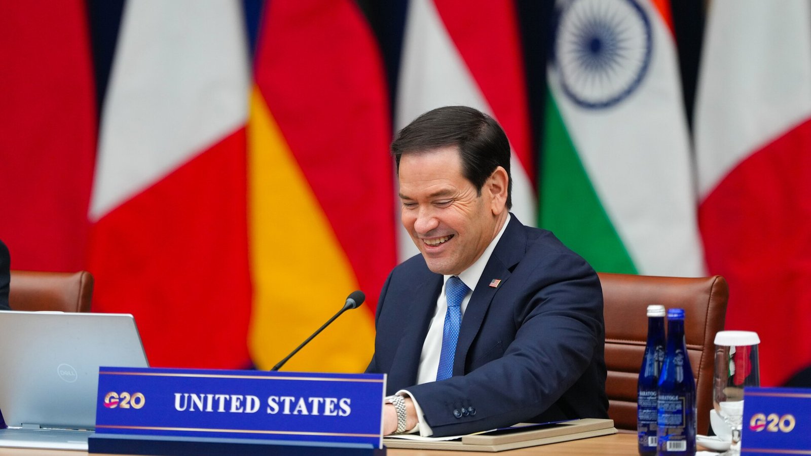 Secretary of State Marco Rubio smiles at the United States seat at the G20 foreign ministers' meeting table, with national flags behind him