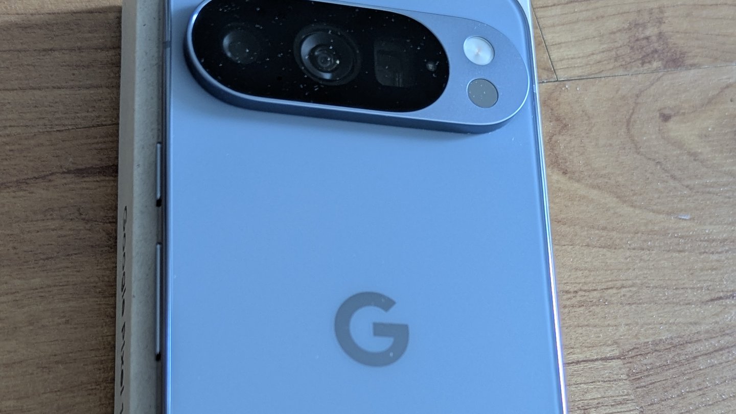 The back of a light blue Google Pixel 10 Pro phone with its camera bar and a grey G logo, lying on a wooden table