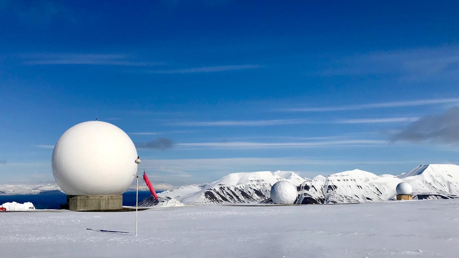 White satellite ground-station domes on a snow-covered plateau at the Svalbard Satellite Station in Norway under a blue sky