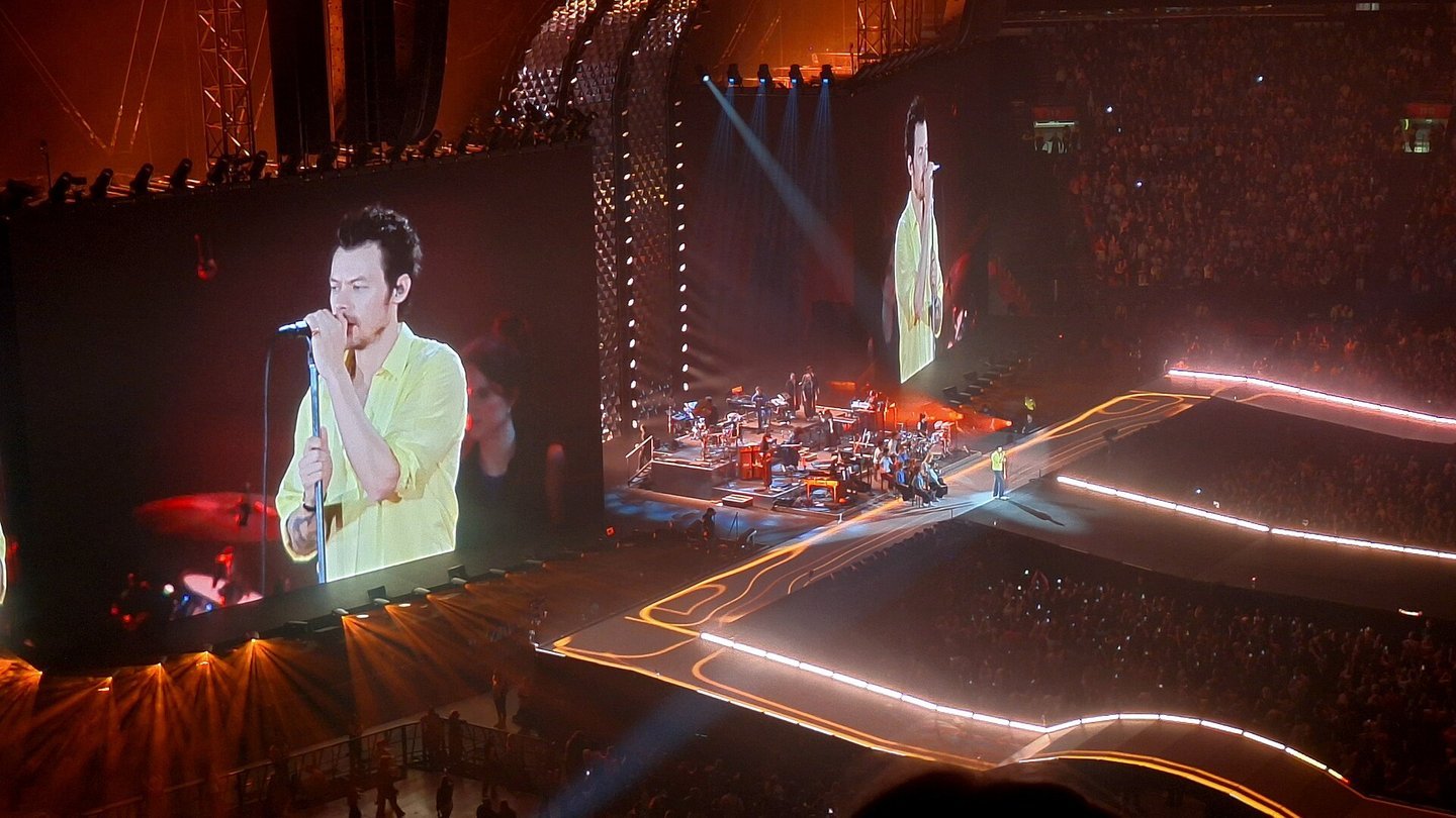 Harry Styles, in a yellow shirt, singing into a microphone on a giant screen above a stage bathed in orange light during a concert in Amsterdam
