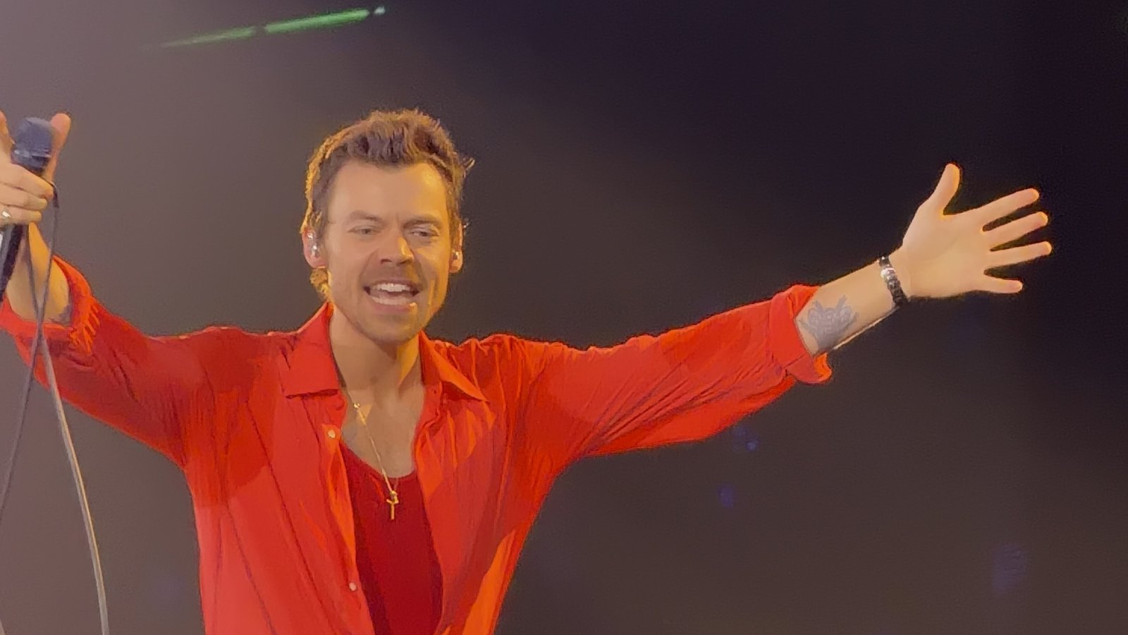 Harry Styles, in a red shirt, holds a microphone and stretches his arms out wide on stage