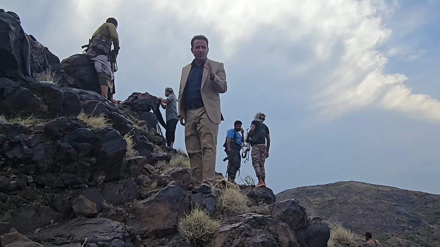 Yemeni government fighters with rifles stand on a rocky hilltop in Jabal Habashi district, Taiz, beside a man in a beige suit