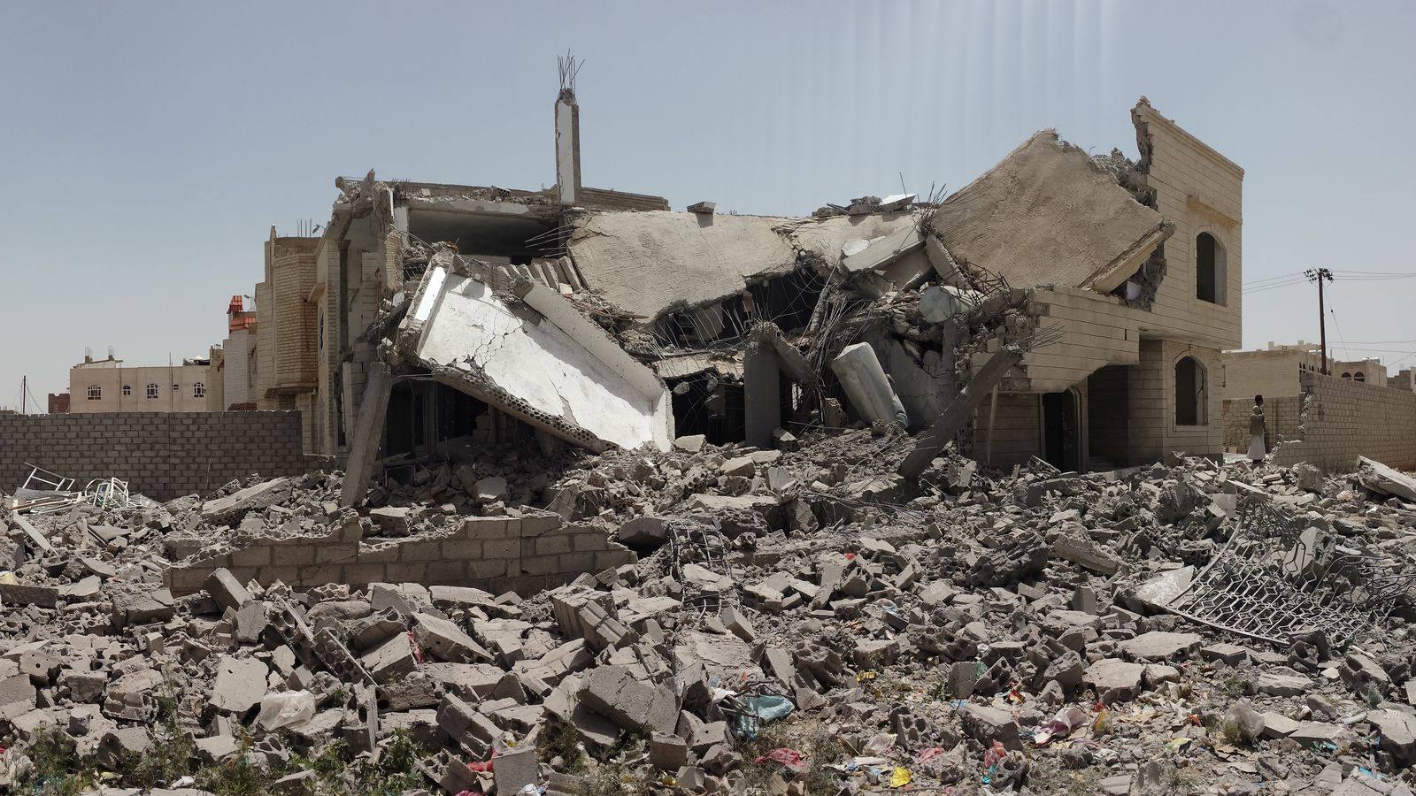 A collapsed multi-storey concrete house surrounded by piles of rubble and broken blocks under a clear sky in southern Sanaa, Yemen