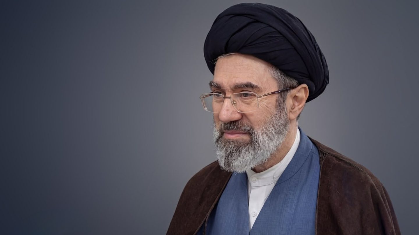 Iran's Supreme Leader Mojtaba Khamenei, wearing a black turban, glasses and a brown cloak, against a grey background