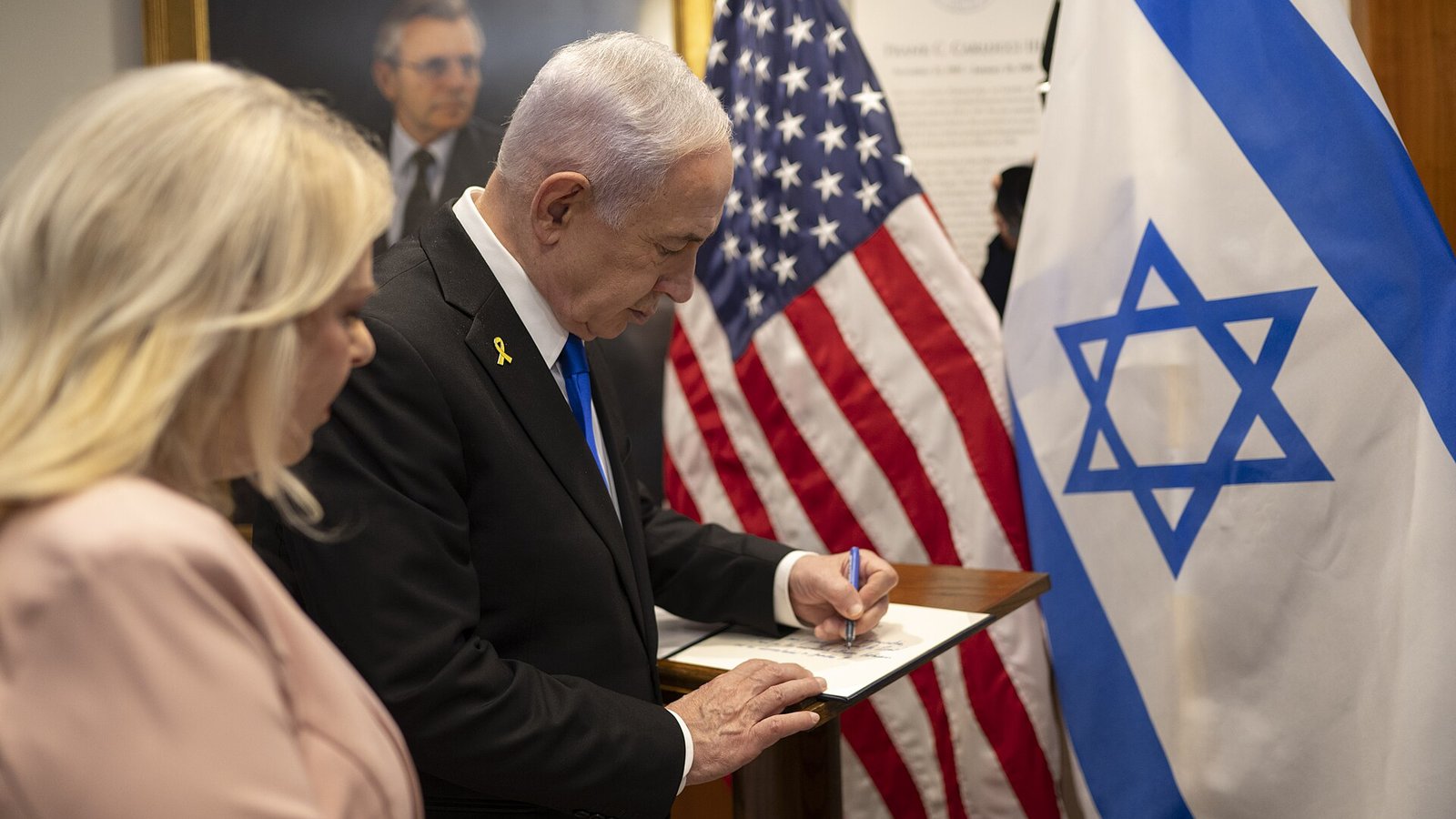 Israeli Prime Minister Benjamin Netanyahu in a dark suit and blue tie signs a guest book at a wooden lectern in front of US and Israeli flags