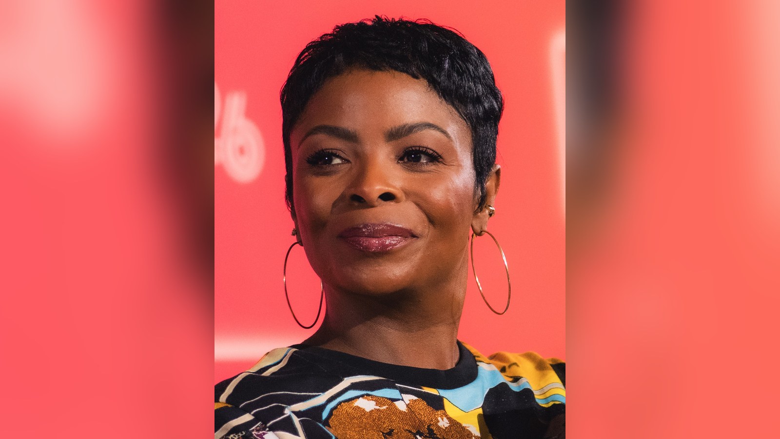 Janelle James, with short dark hair and large gold hoop earrings, smiling in front of a red backdrop