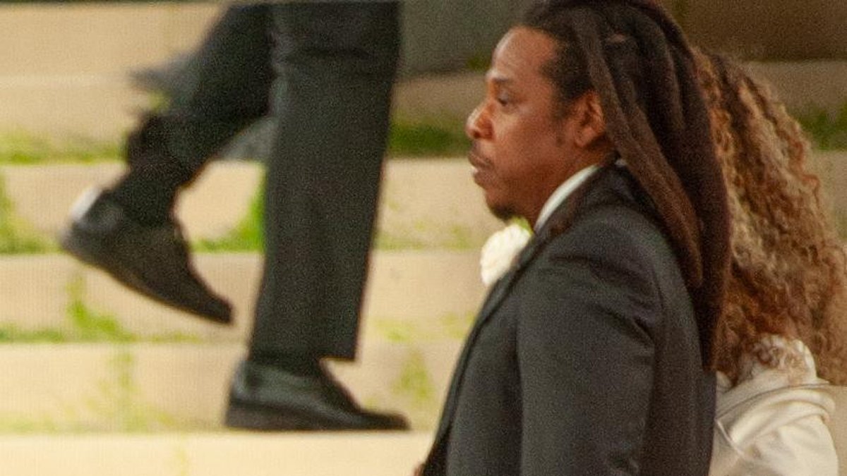 JAY-Z, with long locs and a dark suit with a white flower, seen in profile walking up the steps at the Met Gala