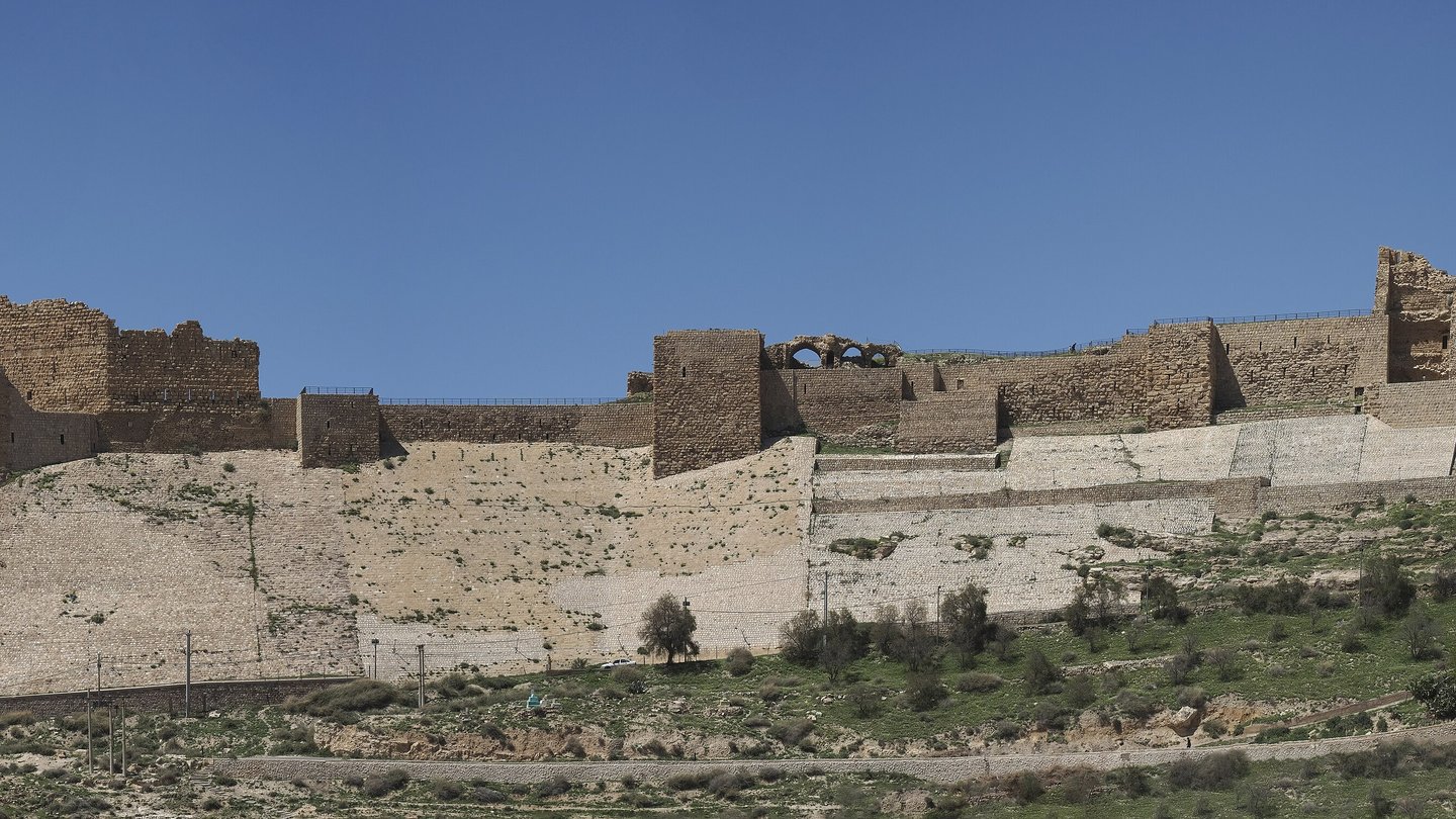 The stone walls of Karak Castle on a hillside under a clear blue sky, with green slopes and houses below
