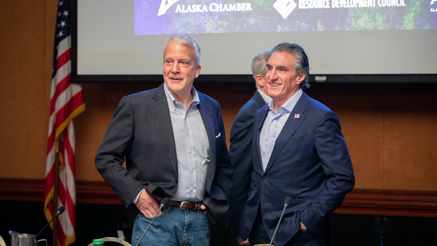 US Senator Dan Sullivan, in a dark jacket and jeans, stands beside Interior Secretary Doug Burgum at a meeting table in Alaska