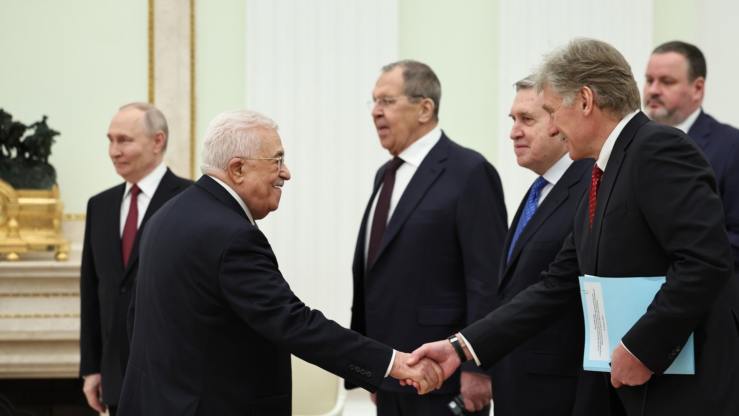 Kremlin spokesman Dmitry Peskov, holding a blue folder, shakes hands with Palestinian President Mahmoud Abbas in a white Kremlin hall as other Russian officials look on