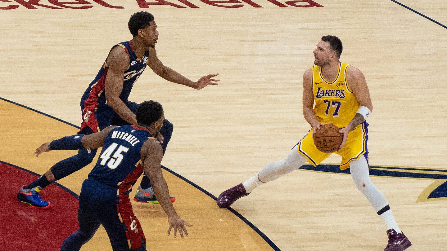 Luka Doncic of the Los Angeles Lakers, in a gold number 77 jersey, holds the ball as two Cleveland Cavaliers defenders guard him