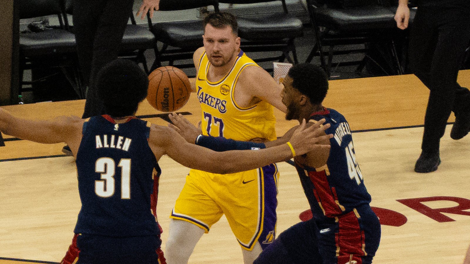 Luka Doncic in a gold Los Angeles Lakers jersey with the number 77 guarded by Jarrett Allen and Donovan Mitchell of the Cleveland Cavaliers