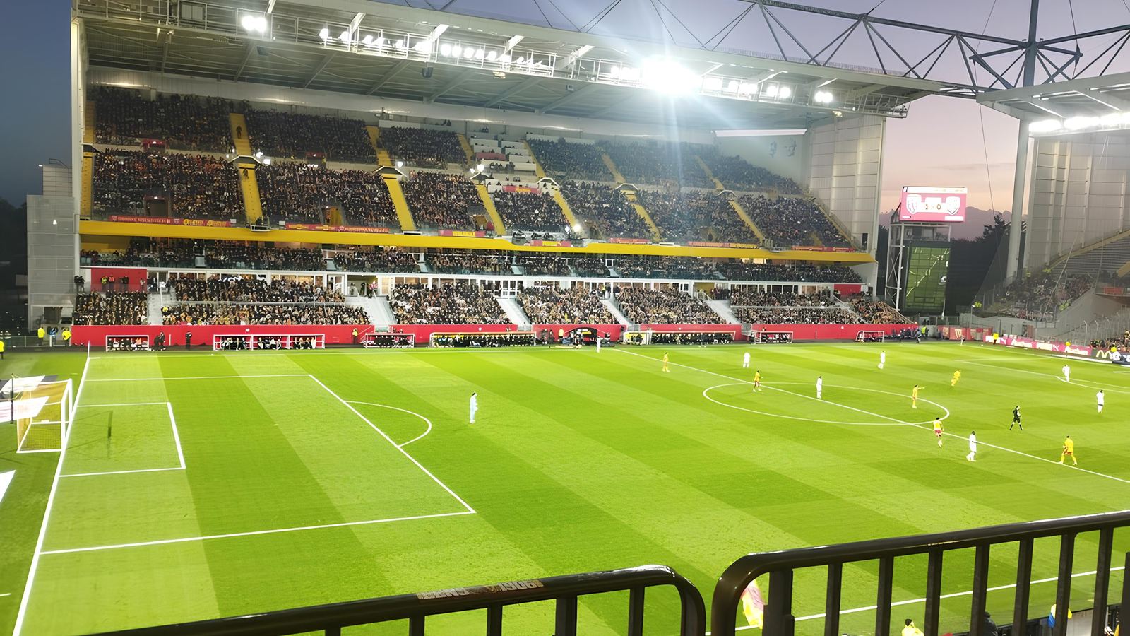 Floodlit evening match at Lens' Stade Bollaert-Delelis seen from the stands, with players on the pitch and a full stand opposite