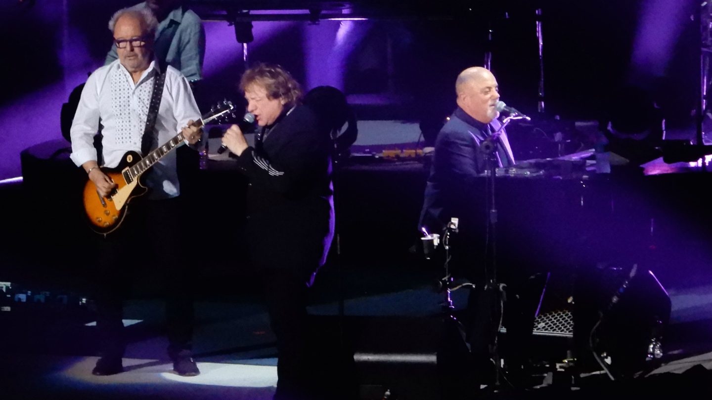 Mick Jones playing guitar, Lou Gramm singing and Billy Joel at the piano on a concert stage lit in purple