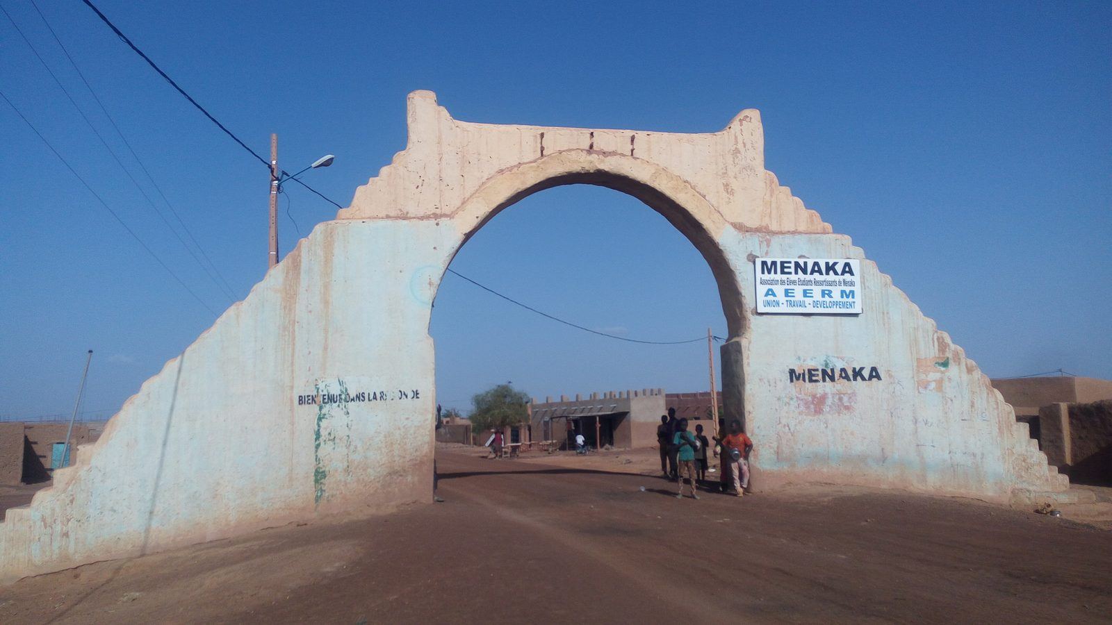 A large whitewashed arch over a dirt road at the entrance to the town of Ménaka in Mali, with signs reading MENAKA, under a clear blue sky