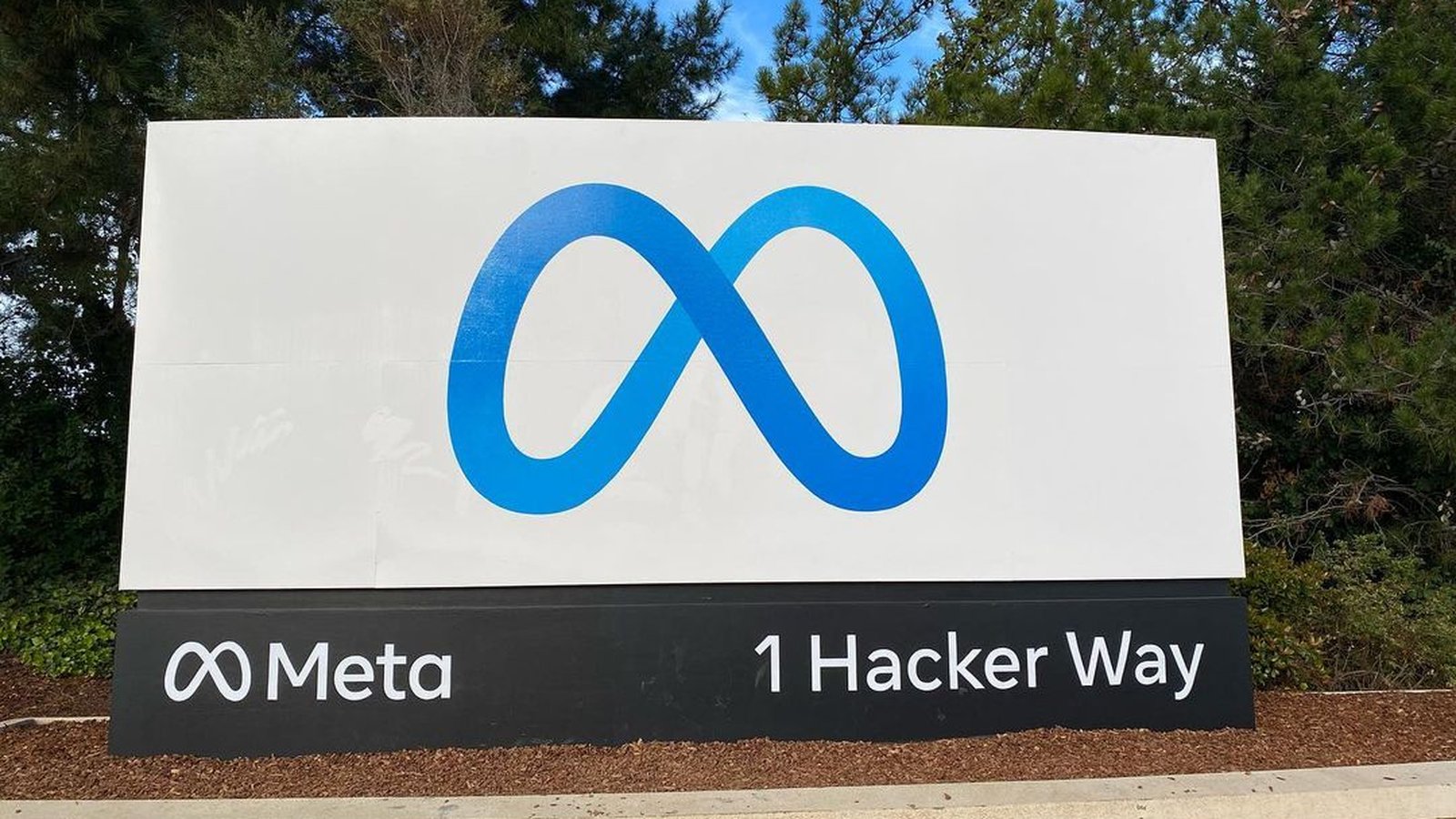A white sign with the blue Meta infinity logo and the address 1 Hacker Way at the entrance to Meta's headquarters, in front of trees