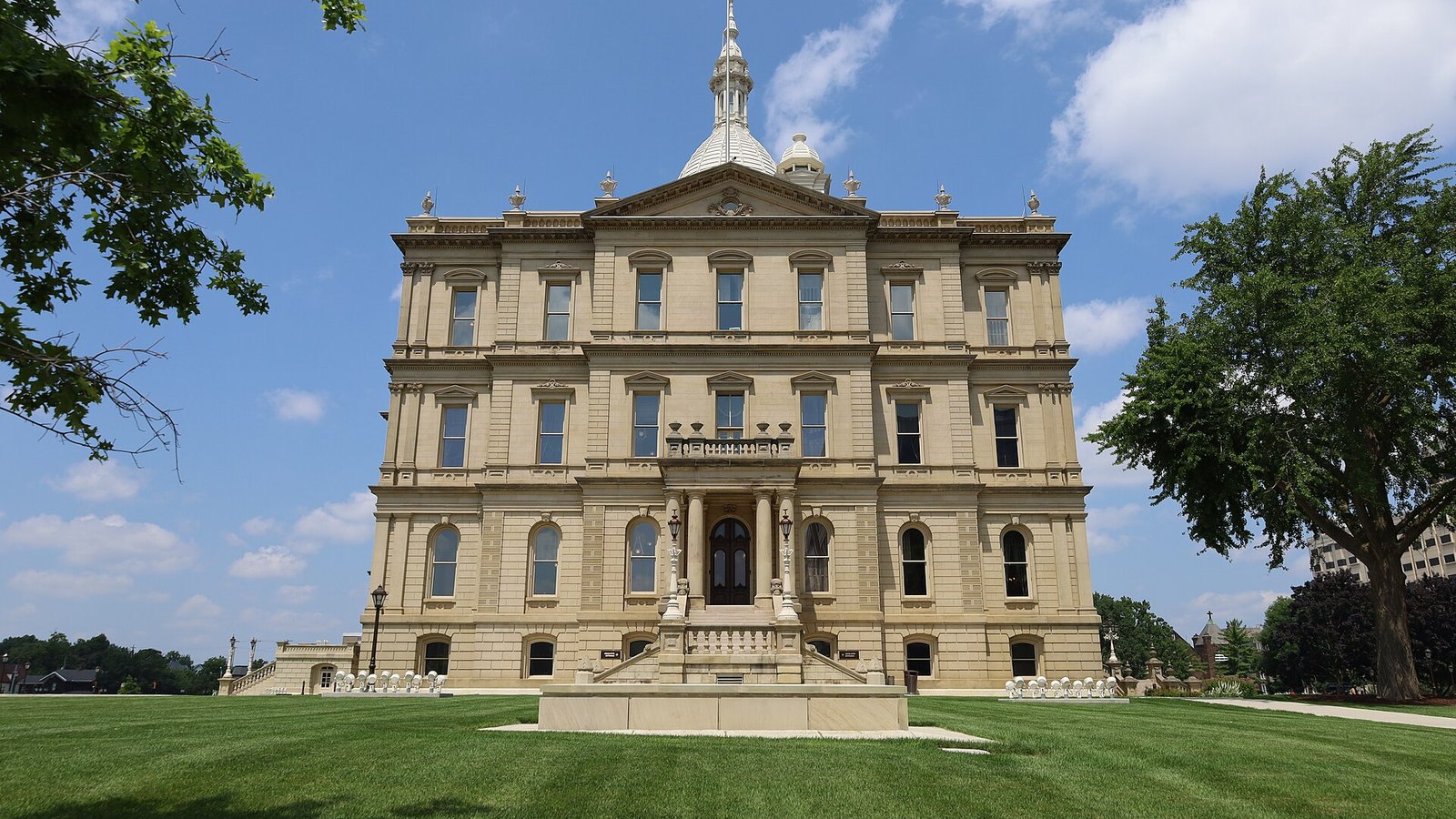 The Michigan State Capitol in Lansing, a domed stone building with a lawn and trees in front, under a blue sky