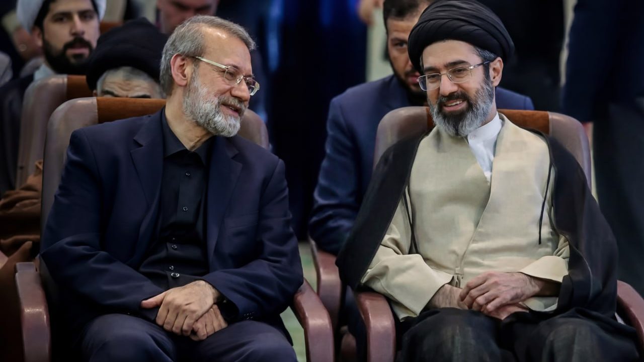 Iran's Supreme Leader Mojtaba Khamenei, in a black turban and cloak, smiles while seated in an audience next to Ali Larijani