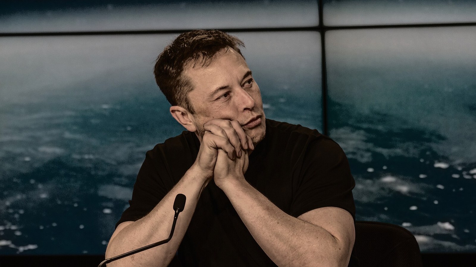 SpaceX and Tesla chief executive Elon Musk sits alone at a press conference desk with a microphone in front of a screen showing Earth from orbit