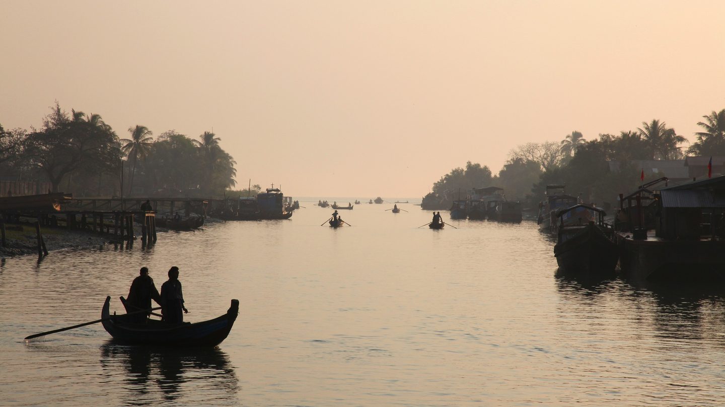 Small wooden boats with rowers on calm water at Sittwe harbour at dawn, with moored boats and palm trees on both banks
