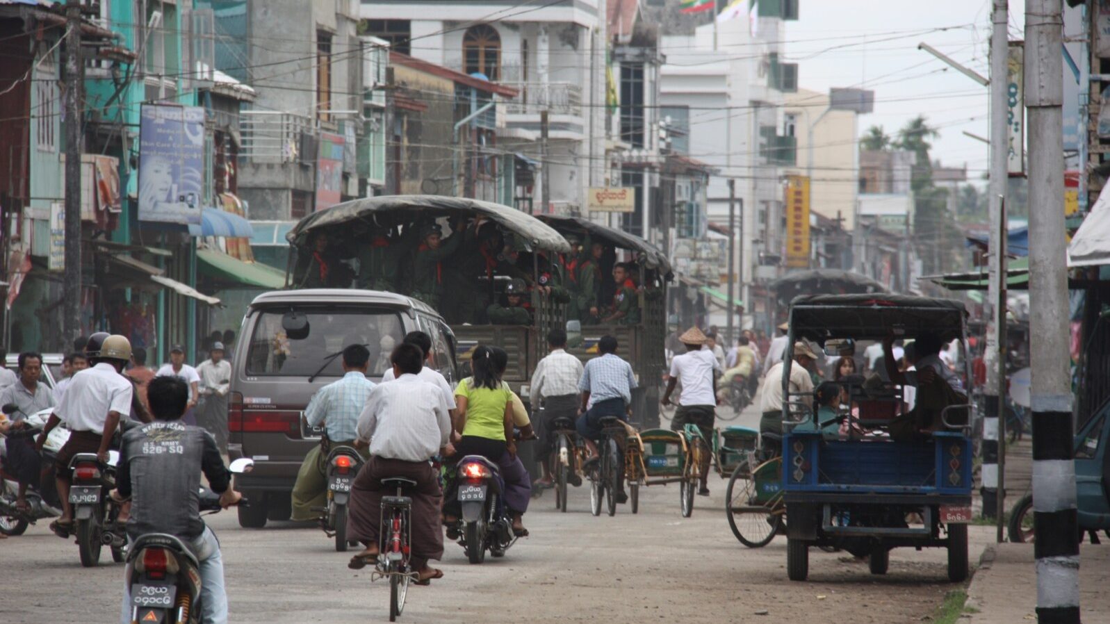 Two military trucks carrying uniformed soldiers drive along a busy street in Sittwe, Myanmar, among motorbikes, bicycles and trishaws