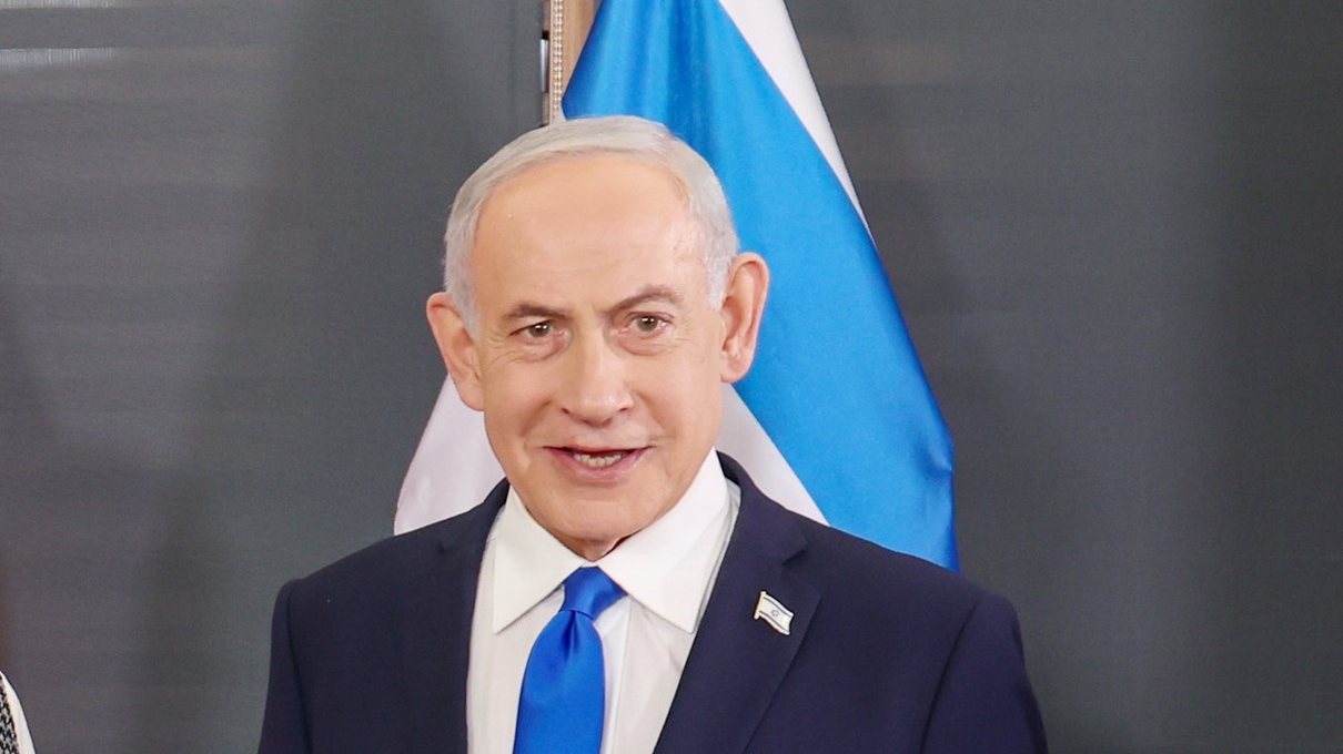 Israeli Prime Minister Benjamin Netanyahu, in a dark suit and blue tie, smiling in front of an Israeli flag