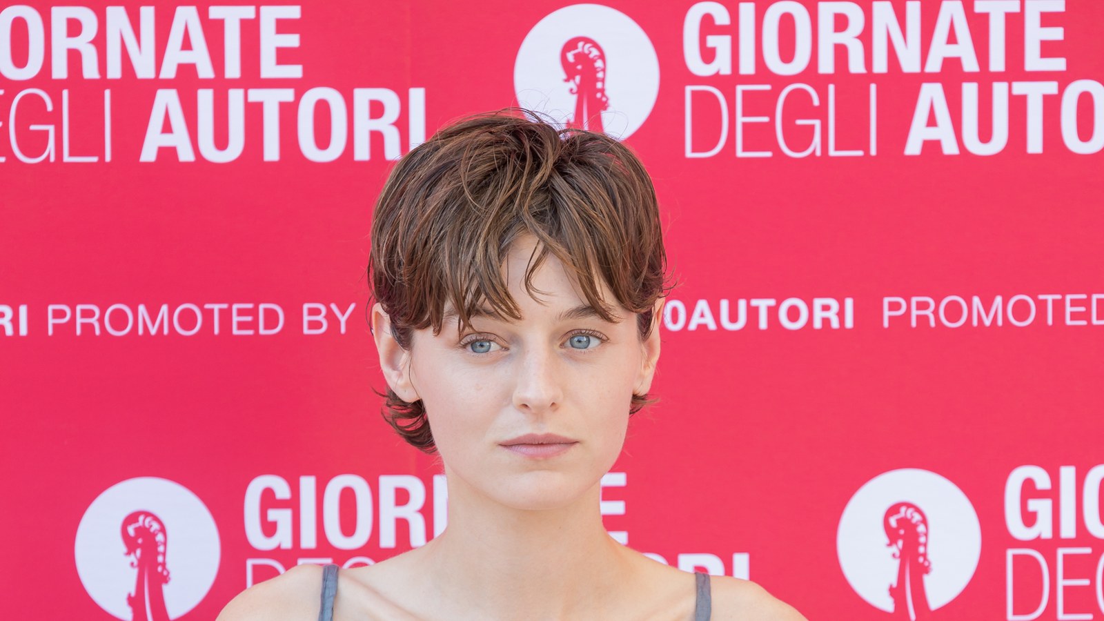 Emma Corrin with short brown hair in front of a red Giornate degli Autori backdrop at the Venice Film Festival