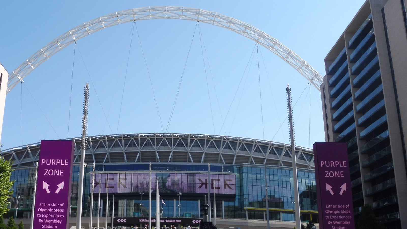 Wembley Stadium's white arch and glass entrance seen from Olympic Way, with concertgoers walking towards the steps